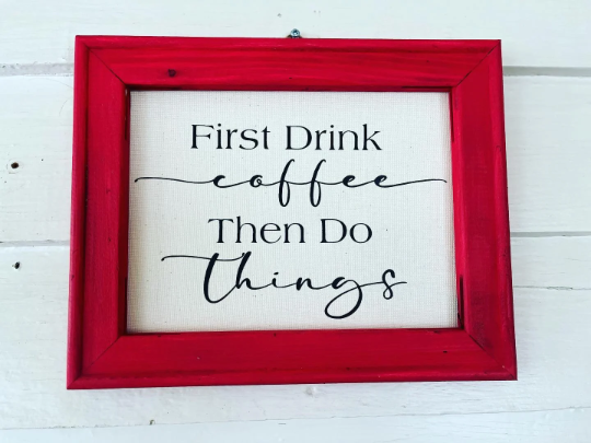 A wooden frame with a canvas wall hanging that reads 'FIRST DRINK COFFEE THEN DO THINGS' in black script, set against a red background.